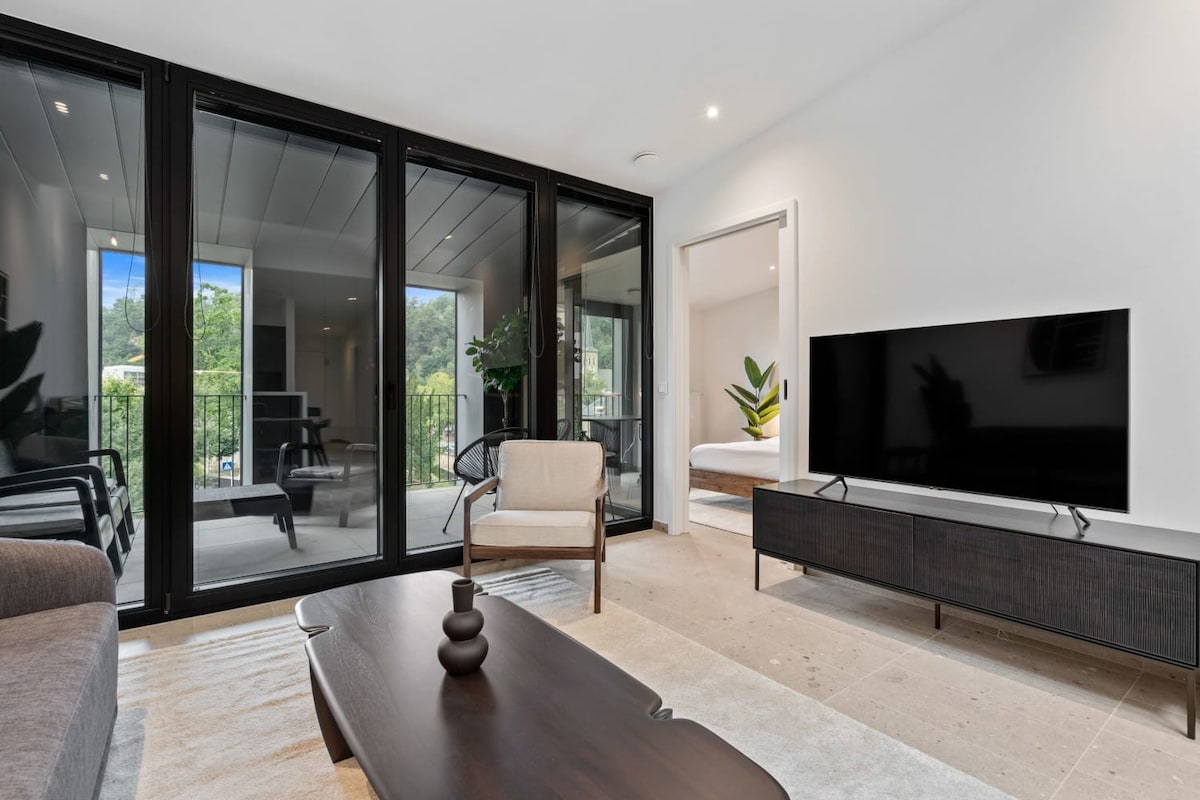 A spacious living area showcases large glass doors that invite natural light and connect to the outdoor balcony. Modern furnishings include a stylish coffee table and a sleek TV stand. Comfortable seating is provided with a minimalistic chair and a plush sofa.