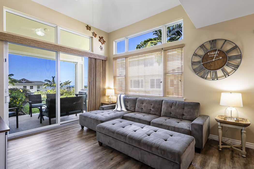 A spacious living area is presented, featuring a large tufted sectional sofa and a coffee table. A wall clock adds character, while large windows allow natural light to fill the space. Sliding glass doors lead to a private lanai with views of the surrounding greenery.