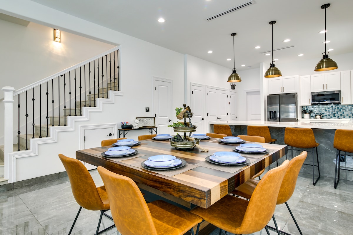 A spacious dining area features a large wooden table set with blue dishware, surrounded by orange leather chairs. The modern kitchen is visible in the background, showcasing white cabinetry and pendant lighting. Stairs lead to the upper level, enhancing the home’s open design.
