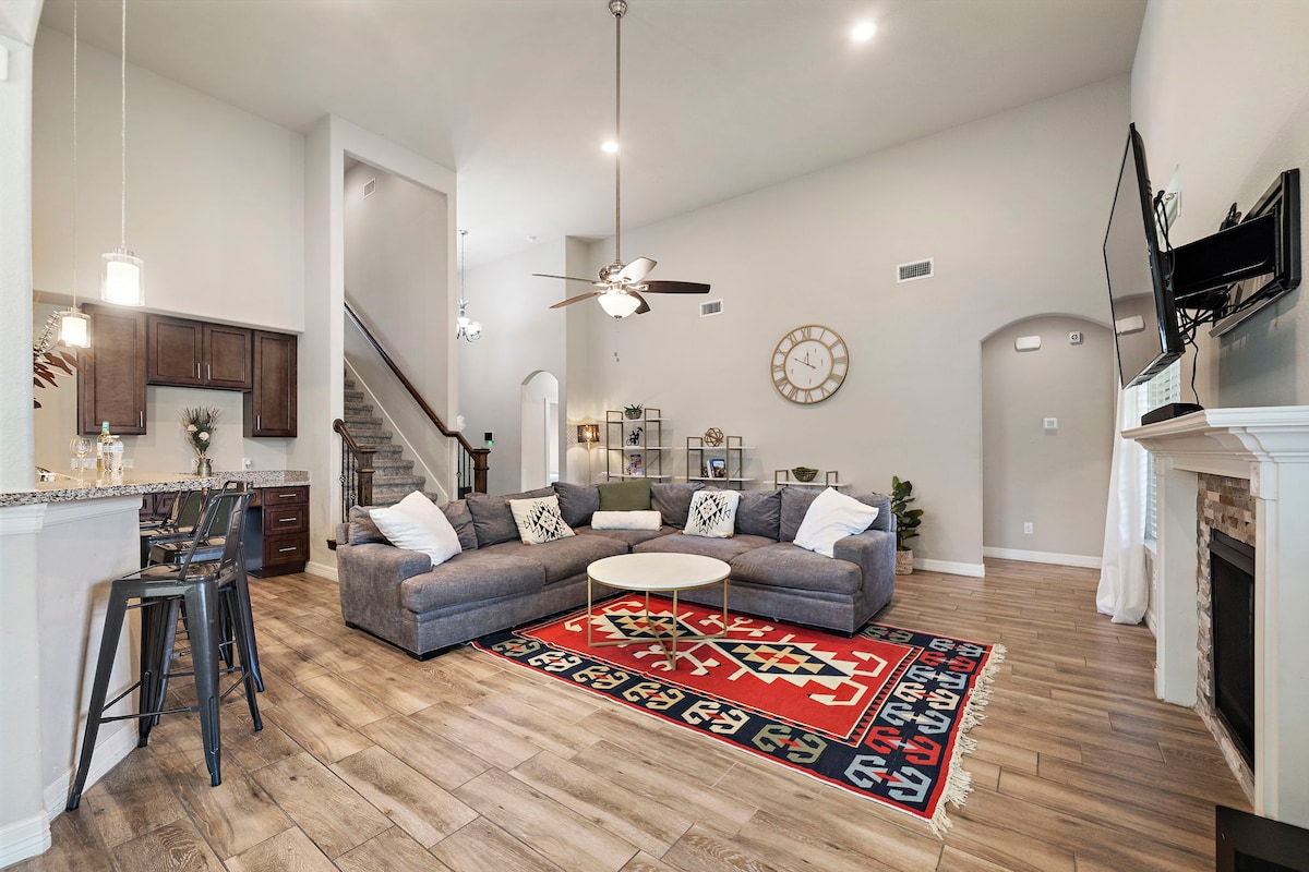 A spacious living area features a large sectional sofa and a circular coffee table atop a vibrant area rug. Bright windows allow natural light to fill the room. The kitchen is adjacent, showcasing dark cabinetry and bar seating. A staircase is visible in the background.