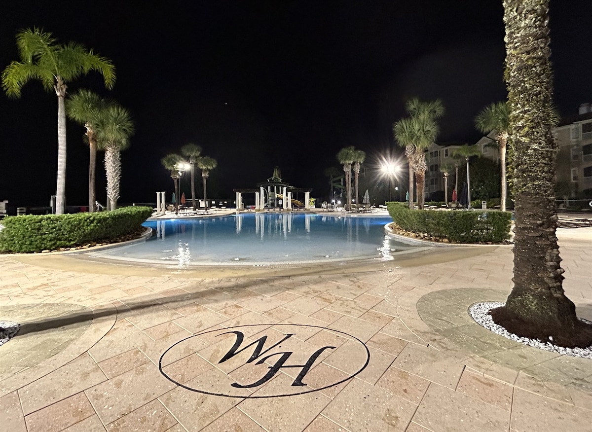A spacious outdoor pool area is surrounded by palm trees and manicured greenery, illuminated by soft lighting at night. The pool reflects the warm glow, with lounge chairs arranged around the perimeter, offering a serene atmosphere for relaxation.