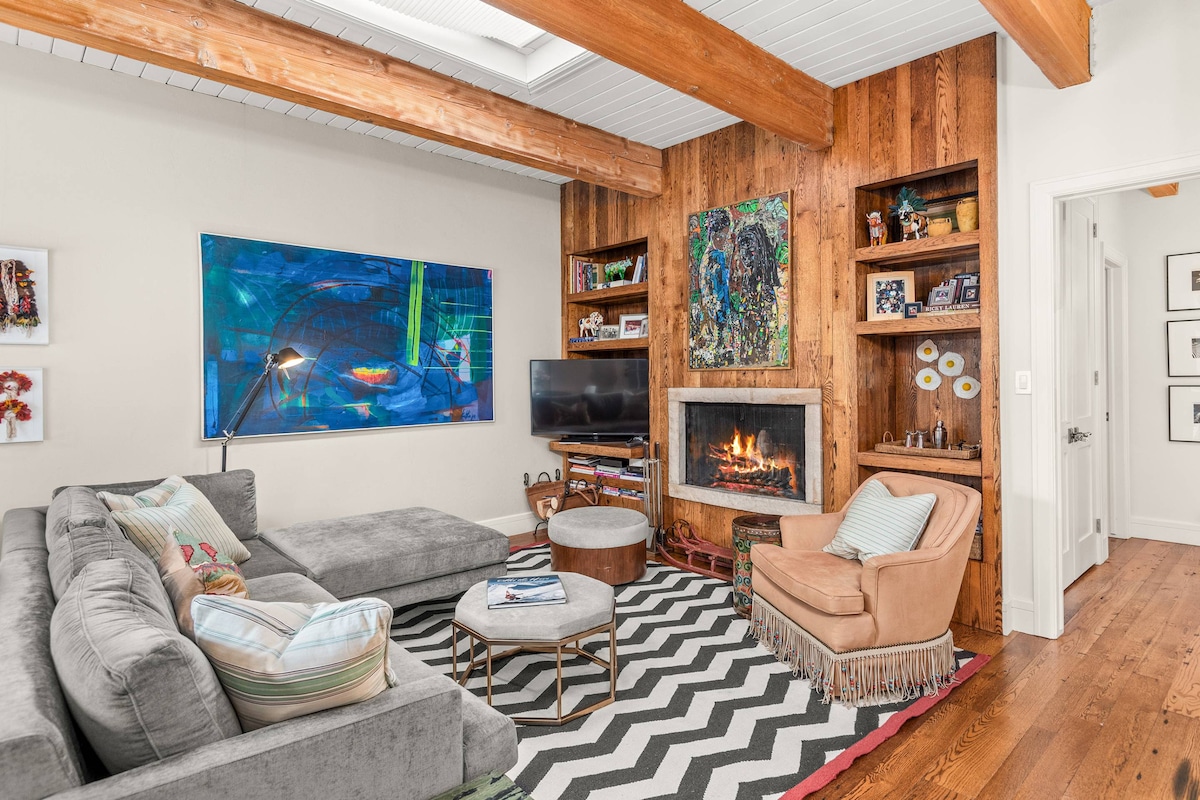 The living room features a sectional gray sofa and an armchair, complemented by a patterned rug. A wall-mounted flat screen TV and a wood-burning fireplace create a cozy focal point. Large windows allow natural light to fill the space, highlighting wooden beams and artwork.