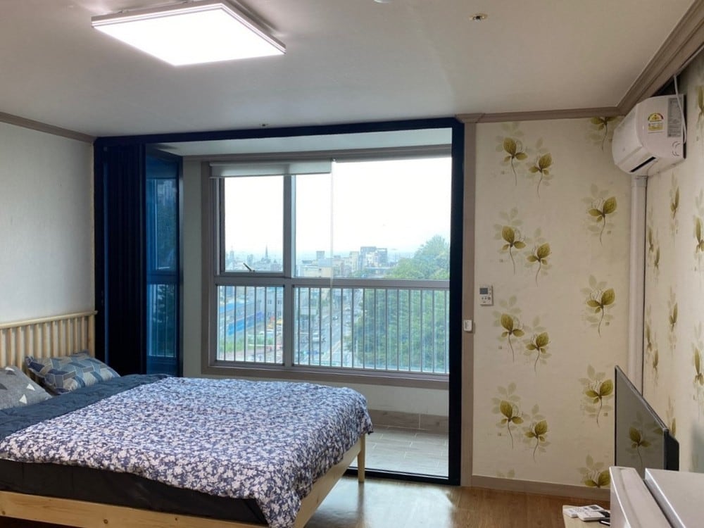 A room features a spacious bed adorned with a patterned blue comforter, complemented by decorative pillows. Large windows provide natural light and showcase an urban view. Floral wallpaper adds a decorative touch, while an air conditioning unit and a television are visible.
