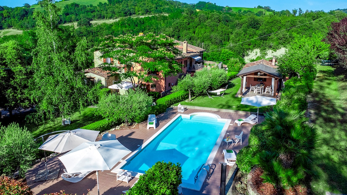 The image showcases a clear blue swimming pool surrounded by loungers and umbrellas. The vibrant green landscape and various trees frame the area, complementing the rustic buildings of the villa in the background. A shaded gazebo offers additional outdoor seating.