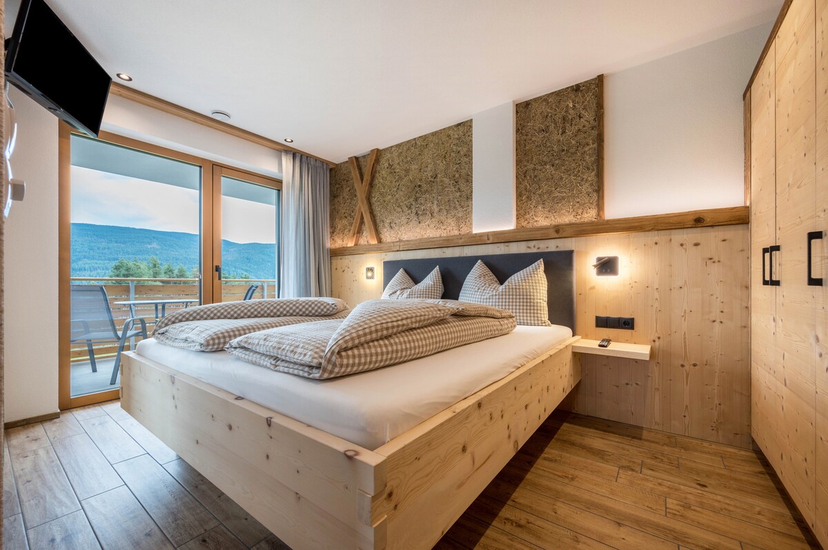 A spacious bedroom features a large wooden bed with textured bedding. Natural light enters through glass doors that lead to a private balcony. Elements of wood and stone accentuate the warm interior, complemented by a mounted television and bedside lighting.