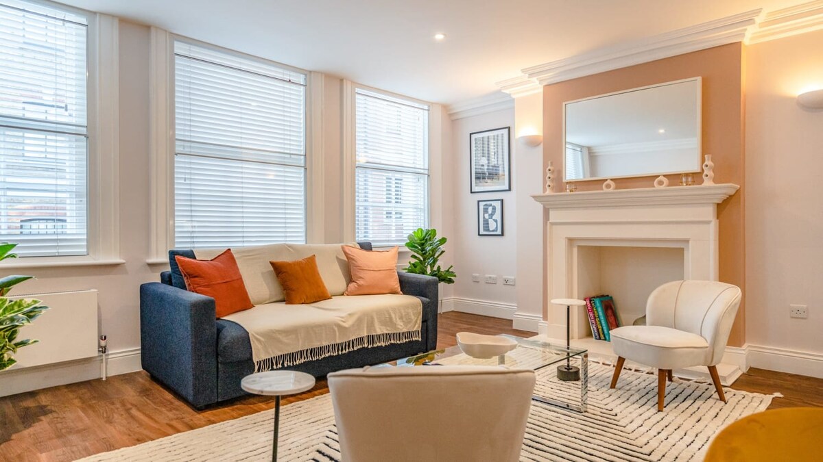 A cozy living area features a sofa adorned with cushions, complemented by a stylish armchair. A light rug covers the wooden floor, while large windows allow natural light to fill the space. Decorative plants enhance the atmosphere, and a mirror reflects the room's brightness.