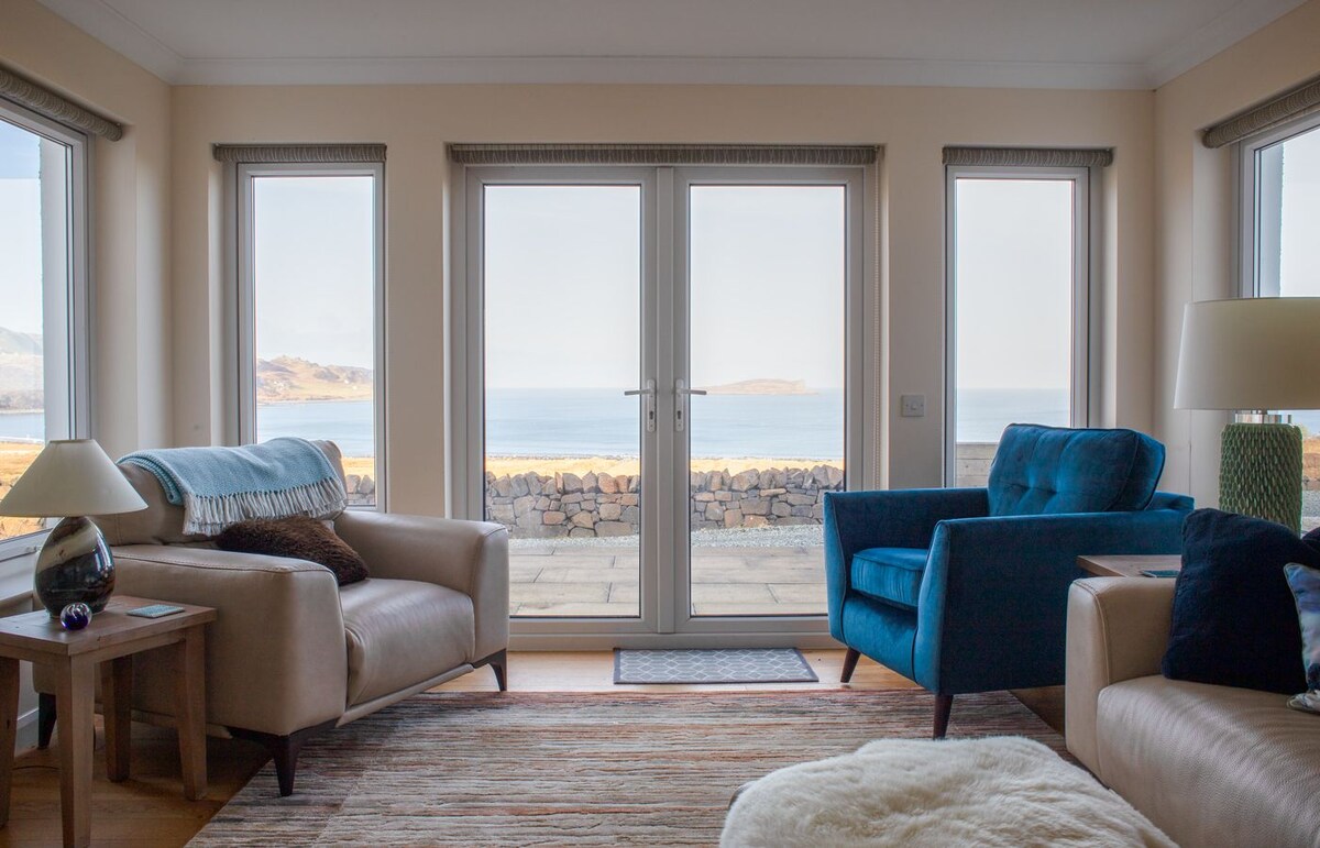 A bright and cozy lounge area features two comfortable sofas, with a textured area rug beneath. Large French doors open to reveal sweeping views of the sea and distant hills. Natural light fills the space, enhancing a sense of relaxation.