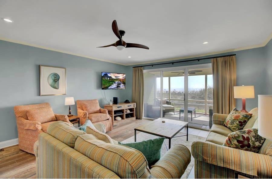 A bright living area features comfortable seating with a striped couch and armchairs. A television is mounted on the wall. Natural light enters through large sliding glass doors, leading to a screen porch. Soft wall colors complement the wooden flooring.