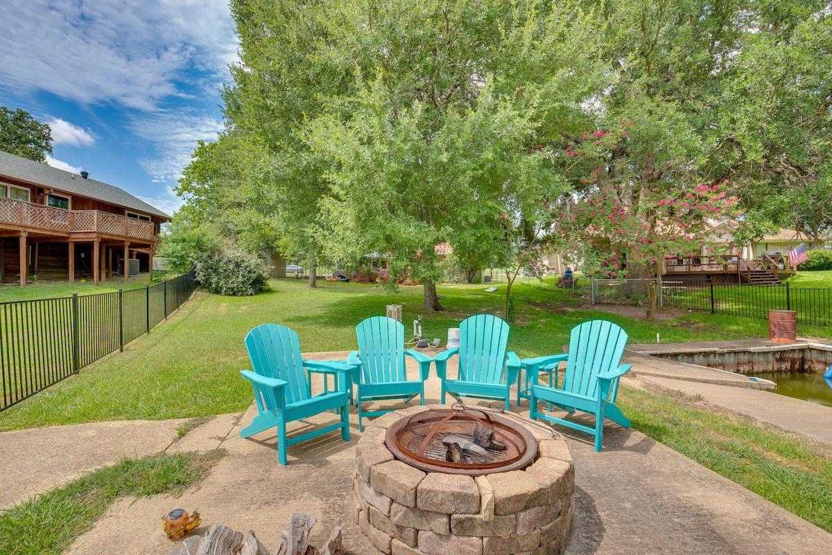 A fire pit area is surrounded by four turquoise Adirondack chairs, set on a concrete patio adjacent to lush green grass. Trees provide a natural setting, while a fence and neighboring structures are visible in the background.