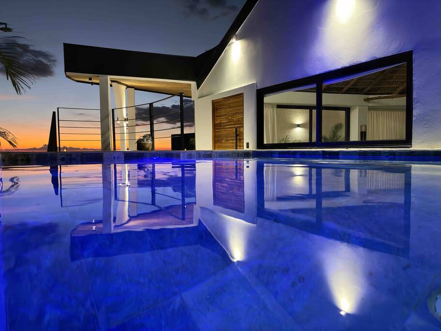 A private pool reflects the evening sky, with warm light from the villa highlighting its clear blue water. The modern villa structure features large windows, allowing a view of the indoor area and the surrounding landscape. The tranquil atmosphere is enhanced by the sunset in the background.