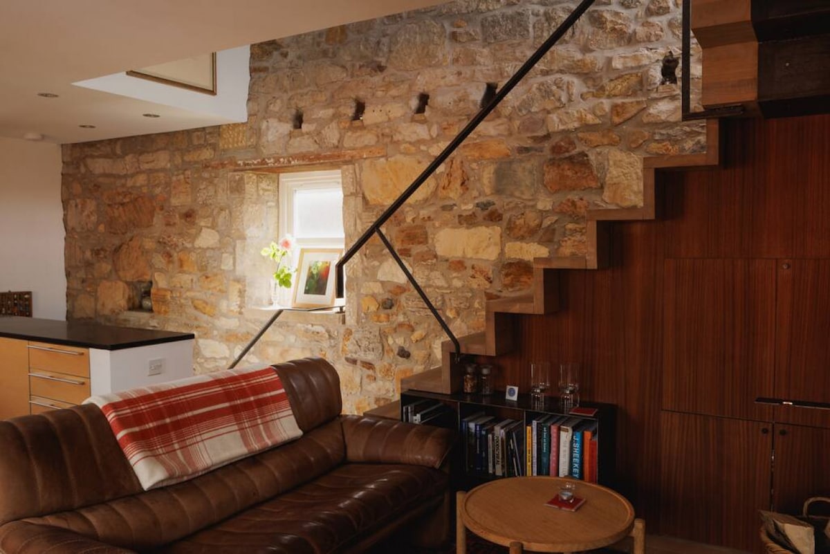An open living area features a blend of stone and wooden elements. A leather sofa is complemented by a plaid throw. A staircase with a sleek railing ascends alongside the exposed stone wall, leading to natural light filtering through a nearby window with potted plants.