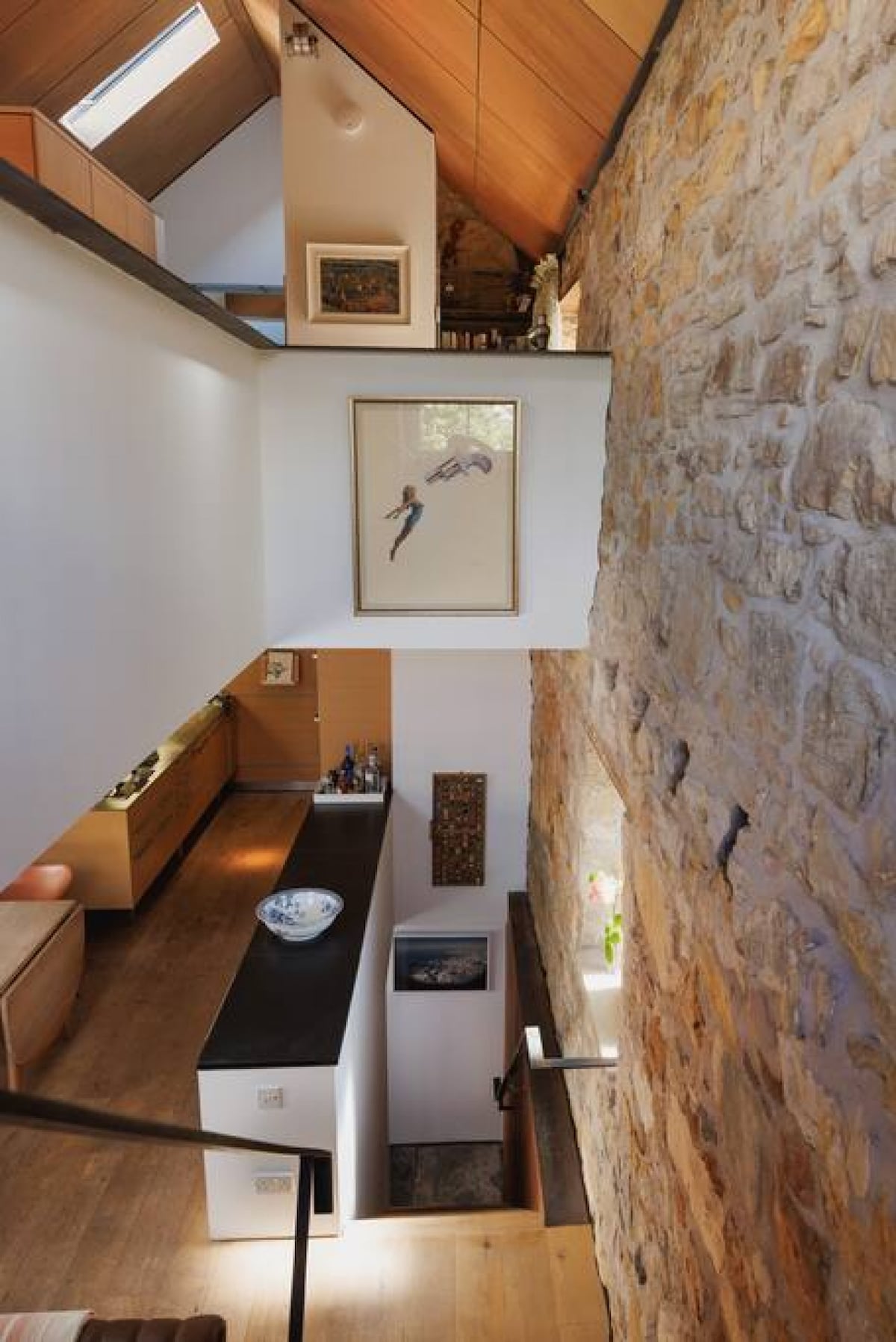 The interior space features a blend of natural stone walls and modern finishes. An artwork is mounted on a white wall, while a wooden ledge holds decorative items. Natural light enters through skylights, enhancing the open atmosphere of the area.