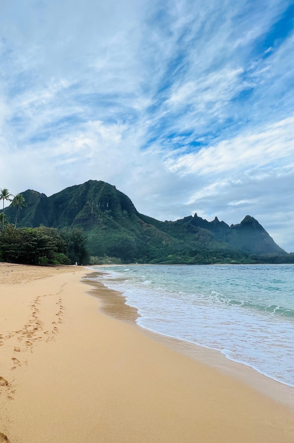 A stretch of sandy beach is bordered by gentle waves lapping at the shore. Towering green mountains rise majestically in the background, accented by a partly cloudy sky. Footprints are visible in the sand, leading towards the water's edge.
