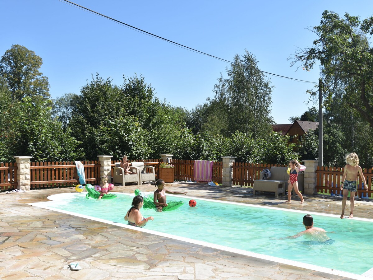 A spacious swimming pool area is depicted, featuring several guests engaged in playful activities. Surrounded by greenery, the area includes comfortable seating and lounge chairs. Colorful pool floats and a beach ball are visible, enhancing the atmosphere of leisure and enjoyment.