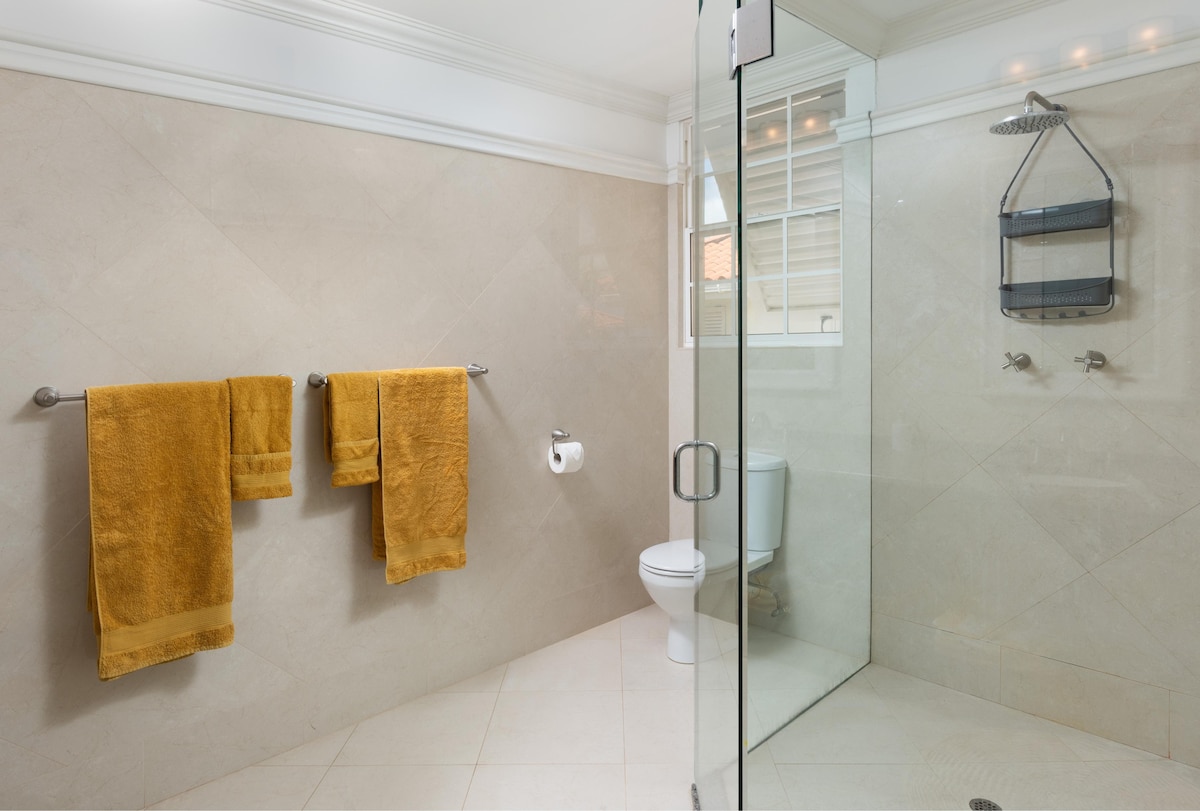 A bright and modern bathroom is highlighted by warm, golden towels hanging neatly on chrome fixtures. A walk-in shower with clear glass panels offers a spacious area, while a white toilet is positioned against the neutral tiled walls.