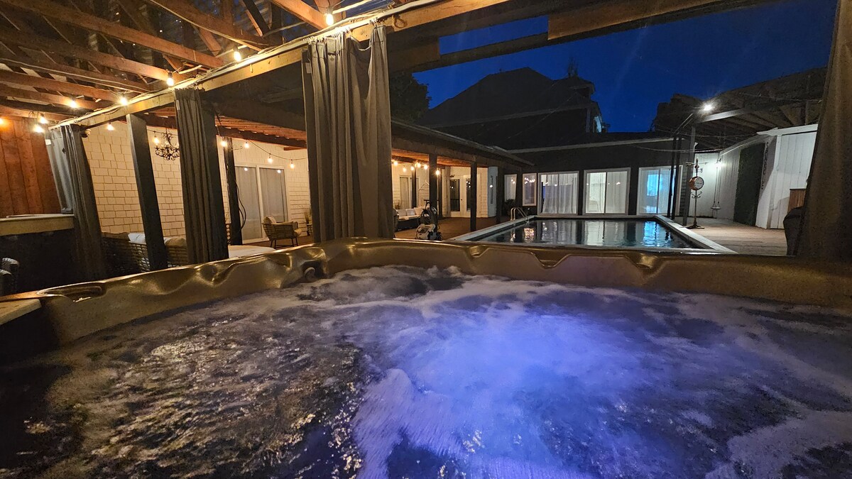 An inflatable hot tub is shown with bubbling water, illuminated in soft blue light under a nighttime sky. The surrounding area features wooden beams and warm string lights. A shared pool is visible in the background, accompanied by lounge seating and a glassed-in porch.