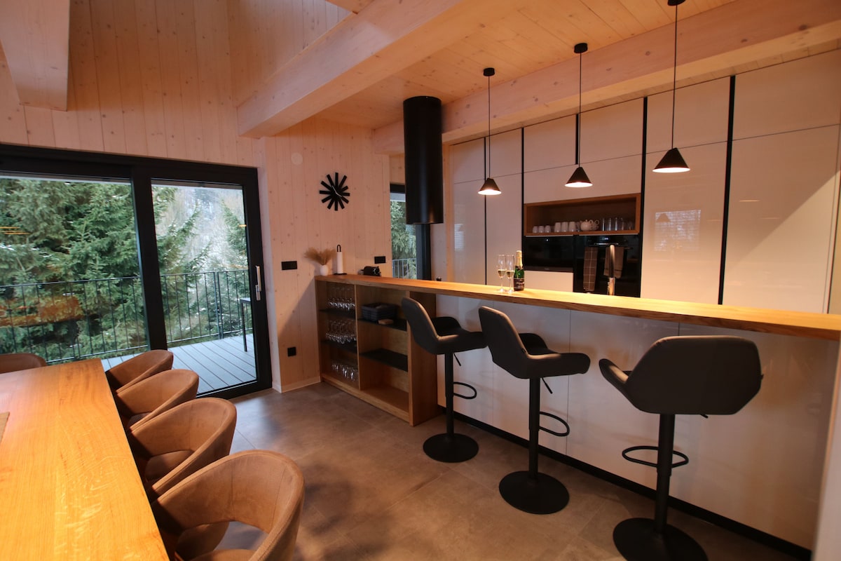 A sleek kitchen space features a large bar with high stools, providing ample seating. Soft lighting from pendant fixtures highlights the minimalist design. Large windows offer views of the surrounding greenery, and the area is complemented by a modern counter equipped with essential appliances.
