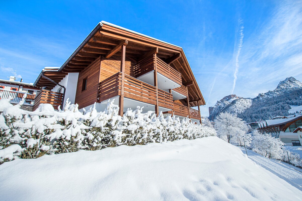 A modern chalet is surrounded by a snow-covered landscape, featuring wooden balconies and a sloping roof. The structure has multiple levels and is set against a backdrop of snow-dusted mountains under a clear blue sky.