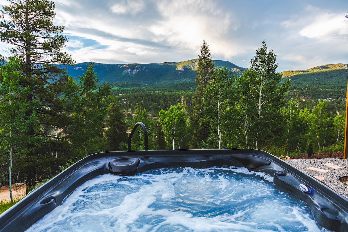 A hot tub, surrounded by trees, provides a serene escape with views of rolling green mountains and a cloudy sky. The bubbling water reflects the natural beauty, inviting relaxation and tranquility in this picturesque outdoor space.