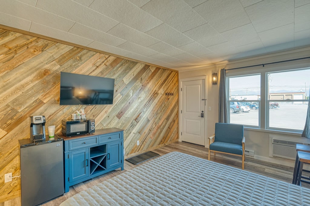 The open-concept living area features a wood accent wall, a 42-inch television mounted above a stylish blue cabinet, which includes a coffee maker and a microwave. A cozy chair is situated near the window, providing a view of the parking area outside.