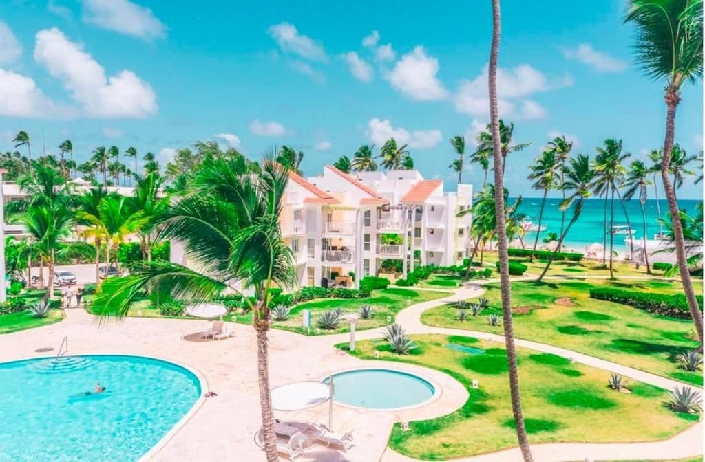 The image displays a vibrant outdoor scene featuring a large swimming pool surrounded by lush green landscaping. White buildings with red rooftops stand in the background, while palm trees sway gently in the breeze. The turquoise ocean is visible in the distance under a bright blue sky.