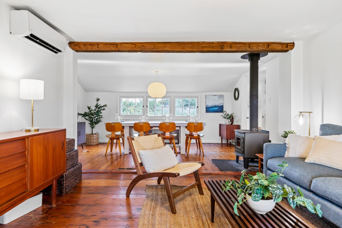 The living and dining area features original wood floors and is illuminated by natural light. A cozy blue sofa, a light wood armchair, and an accent rug complement the space. The dining area showcases a wooden table surrounded by chairs, with a wood-burning stove adding warmth.