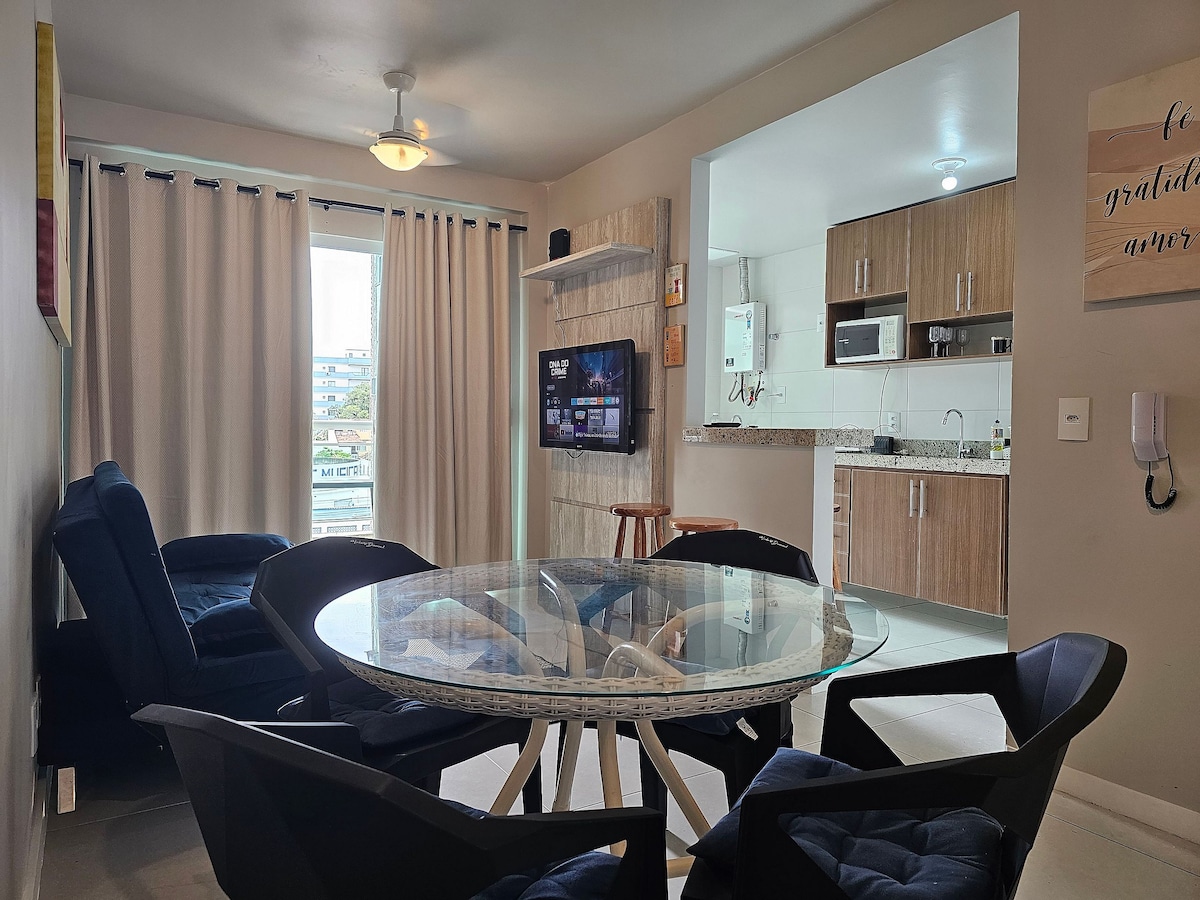 A comfortable living space is featured, with a round glass dining table surrounded by black chairs. A navy blue sofa sits nearby. The kitchen, visible in the background, is equipped with modern cabinets and appliances. Natural light enters through a nearby window, illuminating the area.
