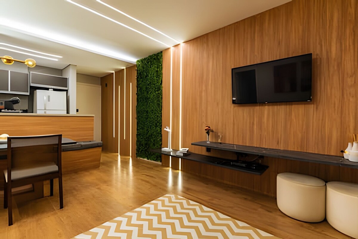 The living area features a modern design with wooden paneling and a green wall accent. A flat-screen television is mounted on the wall above a low, dark console. Comfortable seating is arranged around a dining table, with a textured area rug anchoring the space.