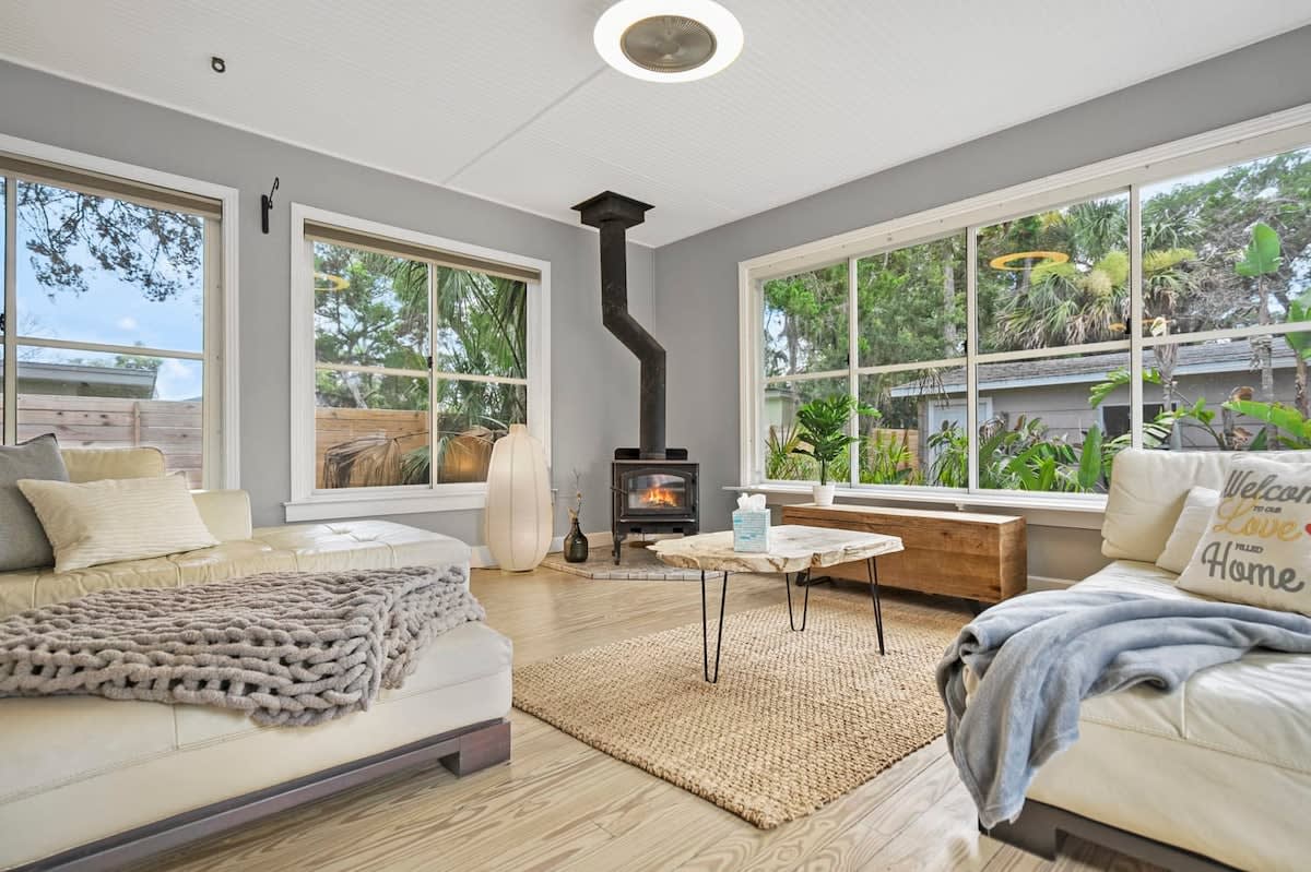 A bright living space features large windows allowing natural light to fill the room. A sleek wood-burning stove stands against a neutral wall. Cozy seating includes a plush white sofa and a light-colored armchair, complemented by a rustic wooden coffee table atop a textured rug.