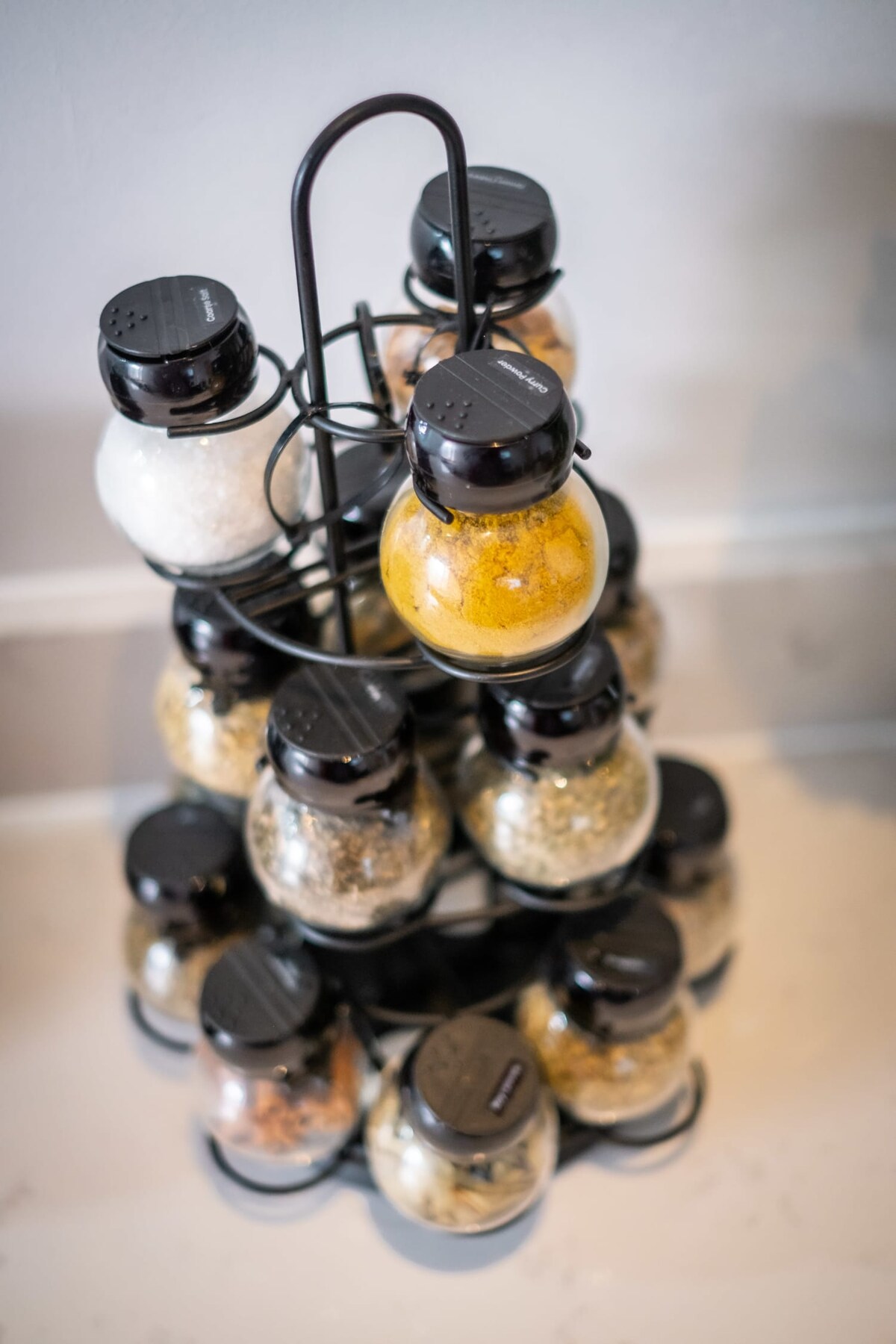 A spice rack is displayed, featuring multiple jars filled with various spices and herbs. The jars are arranged in a tiered metal stand, creating an organized and visually appealing setup. Each jar has a black lid, ensuring freshness and easy access.