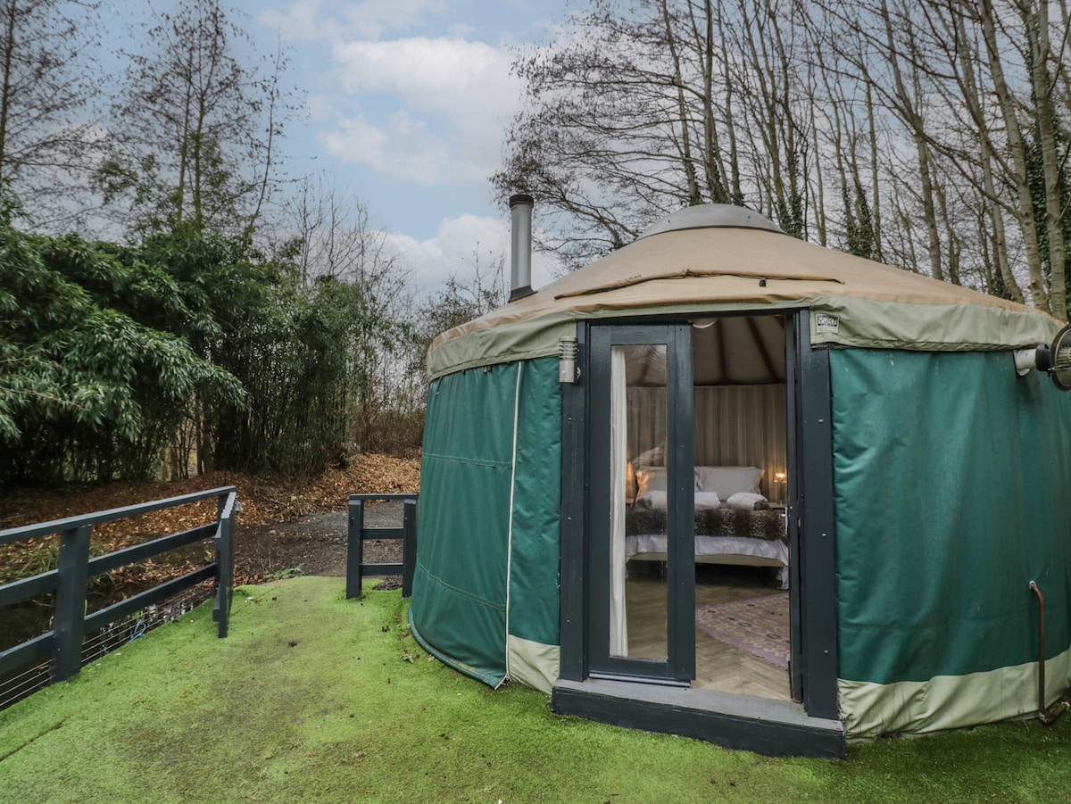 The yurt features a circular design with a green exterior and large windows, providing views of the surrounding greenery. A small wooden veranda leads to the entrance, where soft lighting hints at a cozy interior adorned with comfortable bedding.