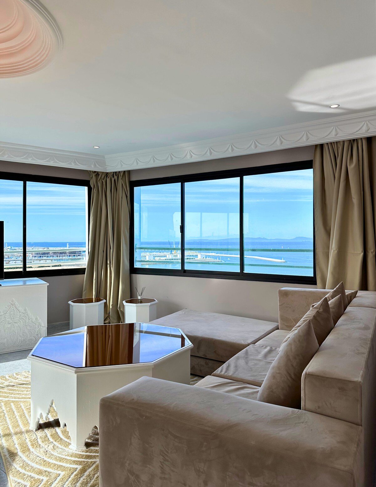 A corner seating area features a light-colored sofa and a glass-topped hexagonal table. Large windows provide expansive views of the water and surrounding landscape, with soft natural light illuminating the neutral decor and light curtains.