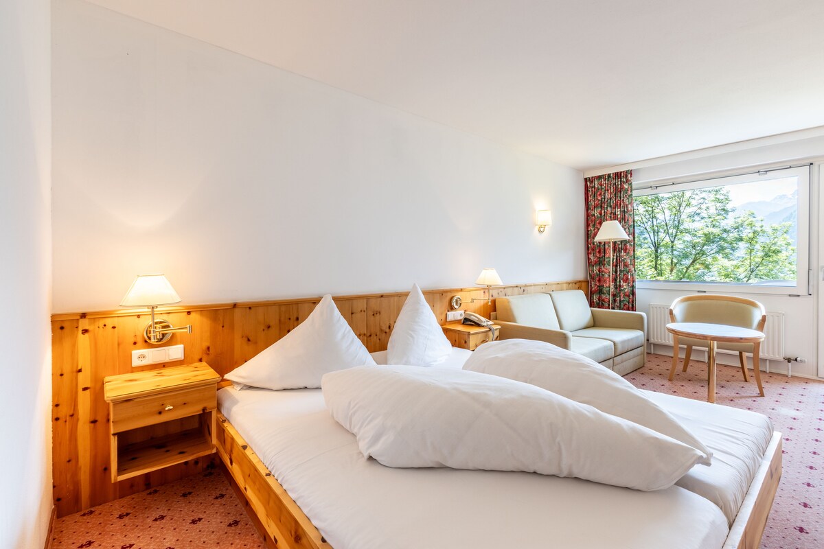 A spacious room features a comfortable double bed with decorative pillows. A seating area includes a light-colored sofa and a round table. Natural light streams in through large windows, offering views of the outdoor greenery and enhancing the tranquil environment.