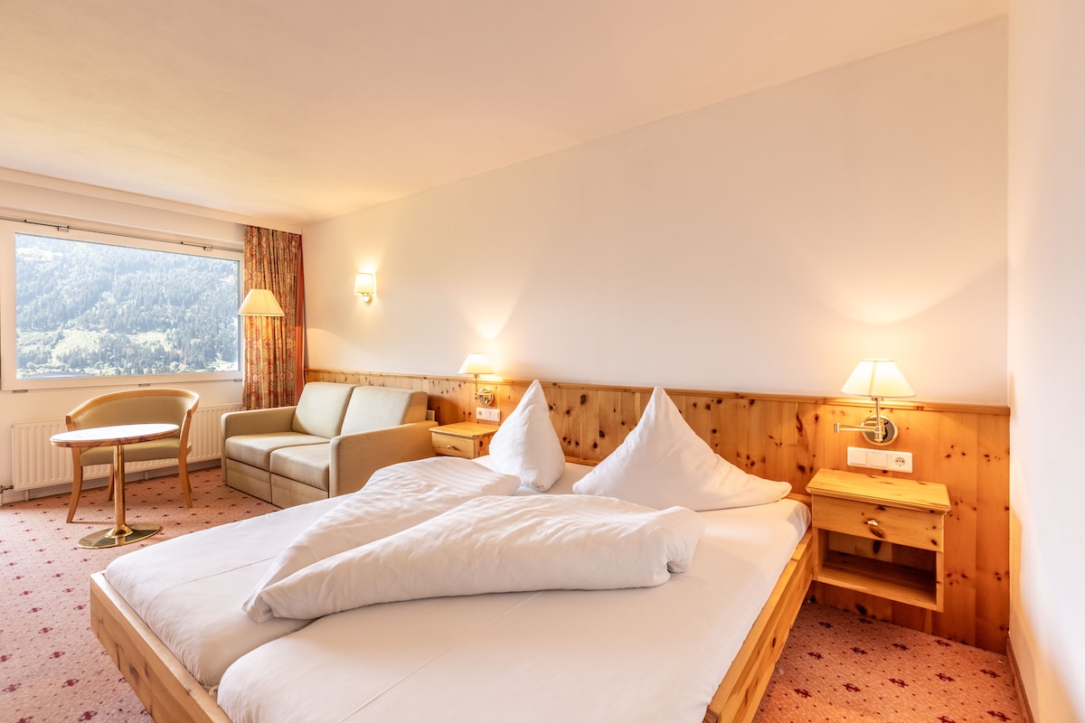 A brightly lit room features a spacious double bed with two decorative pillows. A comfortable seating area is provided with a sofa and a small round table. Large windows offer views of the surrounding mountains, while soft lighting adds to the inviting atmosphere.