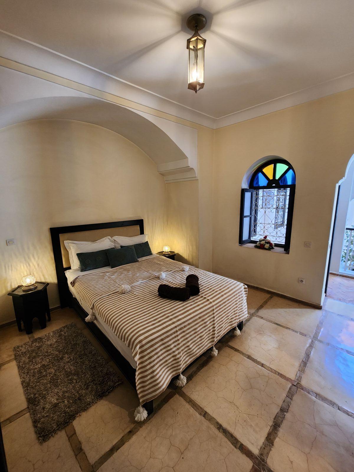 A spacious room features a comfortable bed dressed with neutral-toned linens, complemented by decorative pillows. Soft lighting from bedside lamps creates a calming ambiance. Colorful stained glass windows add character, while polished marble floors provide an elegant finish.