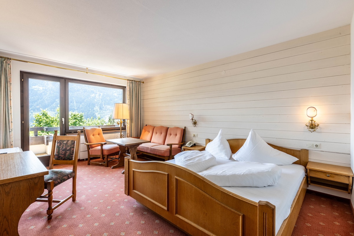 The room features a comfortable double bed with white bedding and two large windows offering panoramic mountain views. A seating area with orange chairs is present beside a wooden table. A desk is located against the wall, providing a functional workspace.
