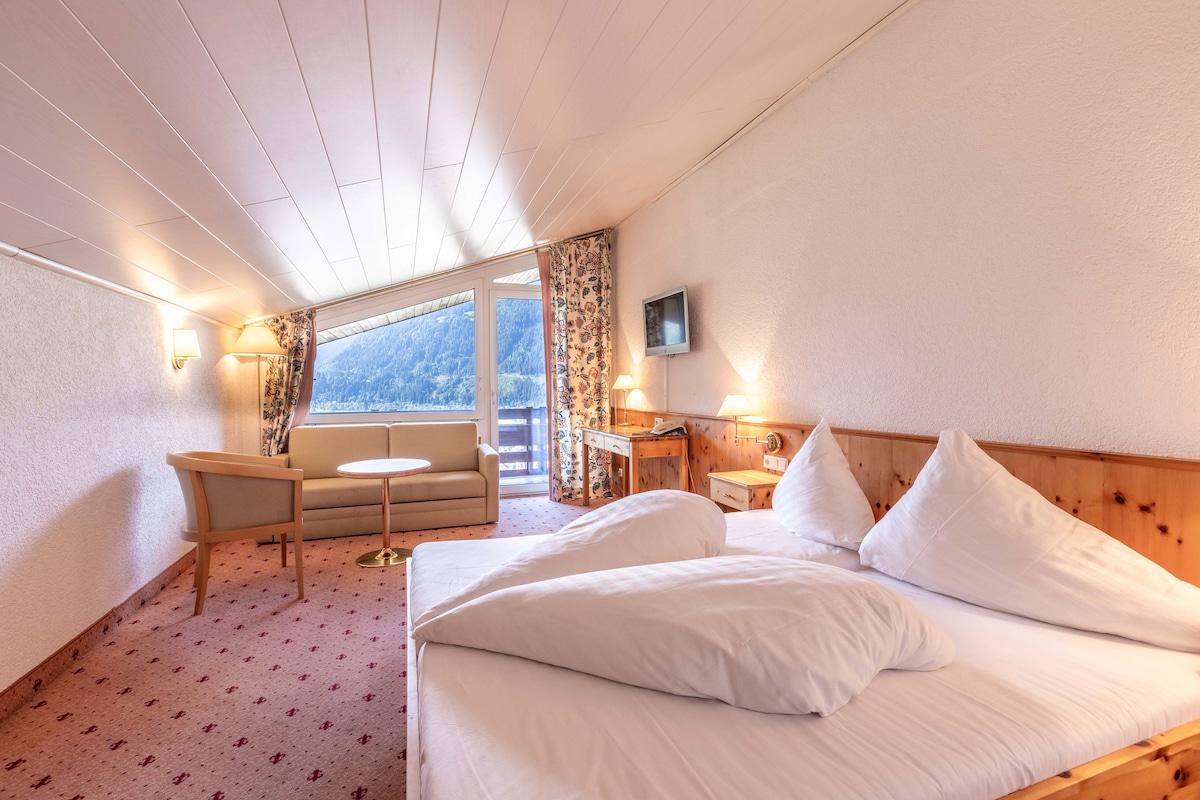 A bright attic room features a cozy double bed with white bedding and pillows. A seating area with a small table and upholstered chairs is present. Large windows provide a panoramic view, complemented by patterned curtains. A flat screen TV and decorative bedside lamps are included.