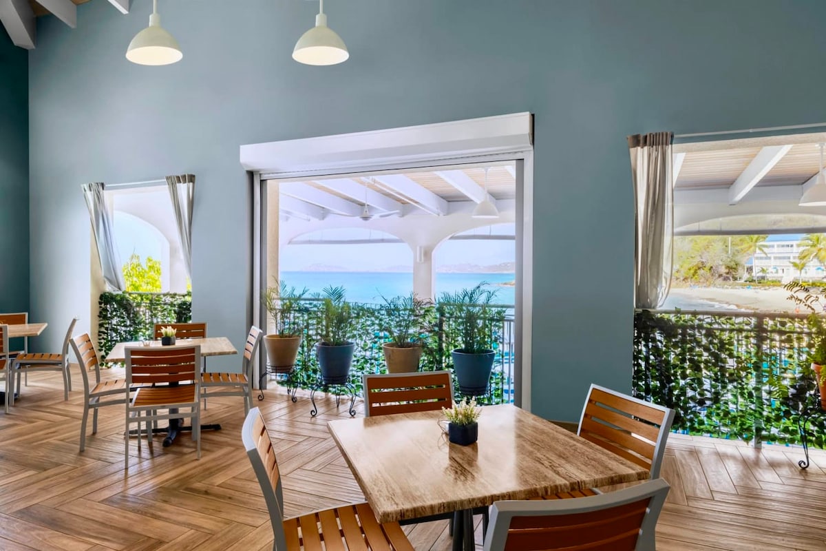 A dining area features several wooden tables surrounded by chairs, with large windows offering views of the beach. Potted plants enhance the ambiance, while natural light floods the space through the open window treatments.