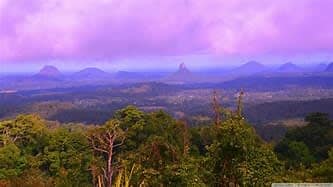 A panoramic view of rolling green hills and distant mountains under a soft purple sky. Lush trees in the foreground frame the landscape, highlighting the natural beauty of the Sunshine Coast Hinterland.
