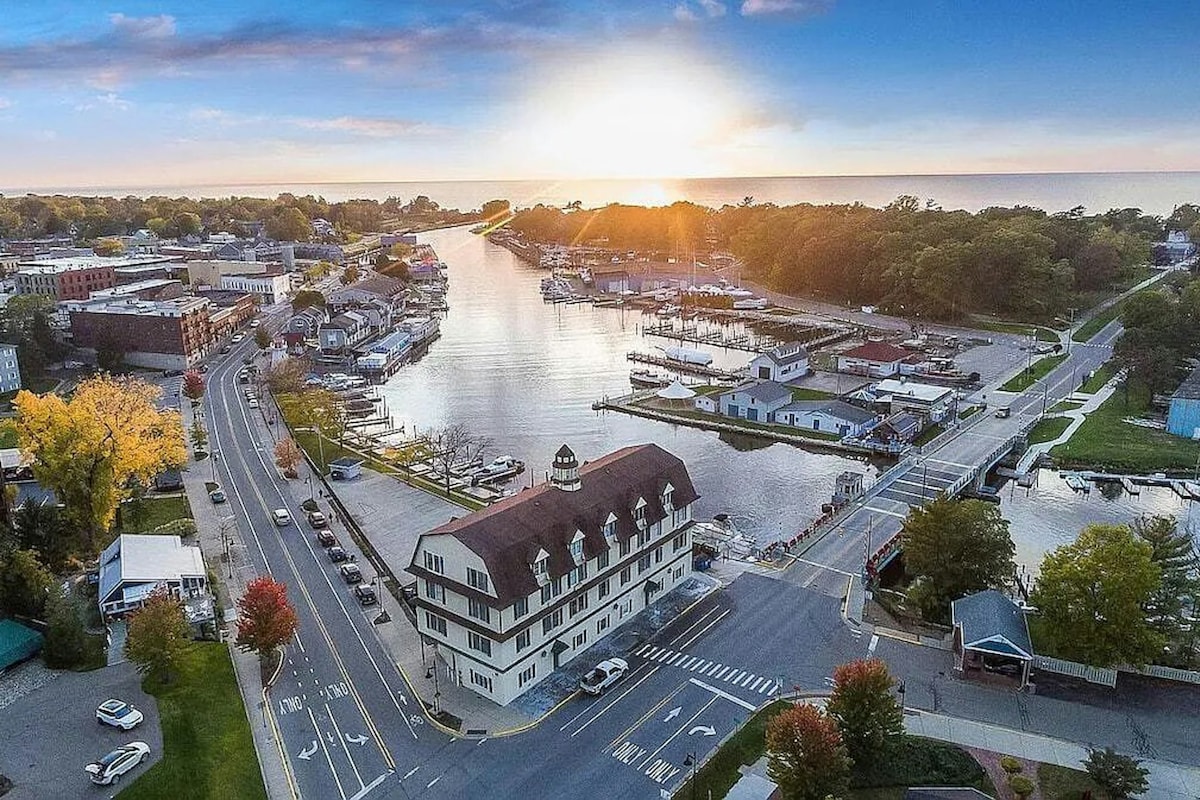 An aerial view captures the winding Black River as it flows into Lake Michigan, surrounded by the vibrant hues of local trees. The buildings along the water are showcased under a sunset sky, reflecting calmness and natural beauty.
