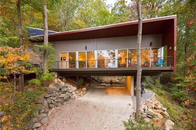 A modern house is perched among trees, supported by a stone and concrete foundation. Large glass windows dominate the front, allowing natural light to fill the interior. The forest surroundings display vibrant autumn foliage, enhancing the connection to nature.