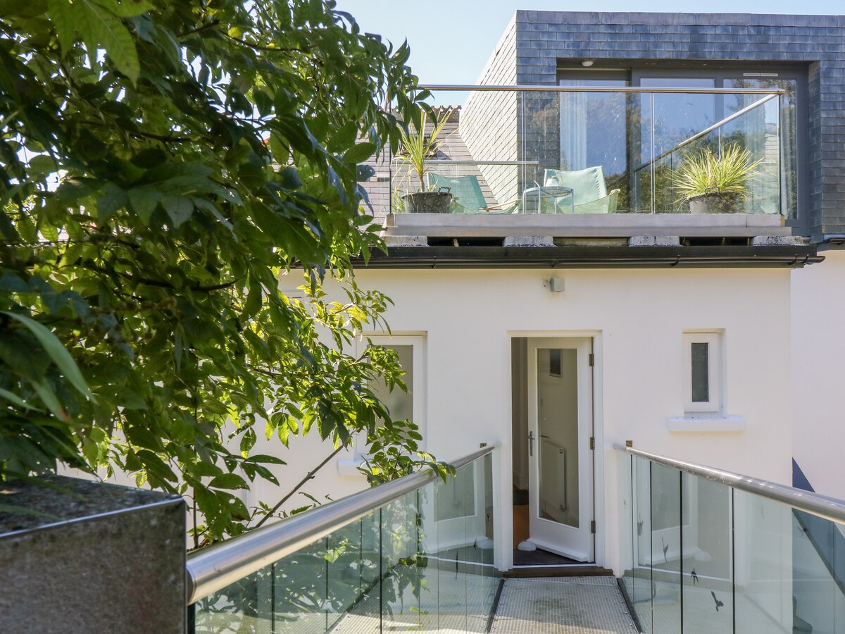 An exterior view showcases a modern duplex with an entrance door and large glass windows. A private bridge leads to a balcony with seating and plans, surrounded by greenery. The building features a blend of light-colored walls and dark slate accents.