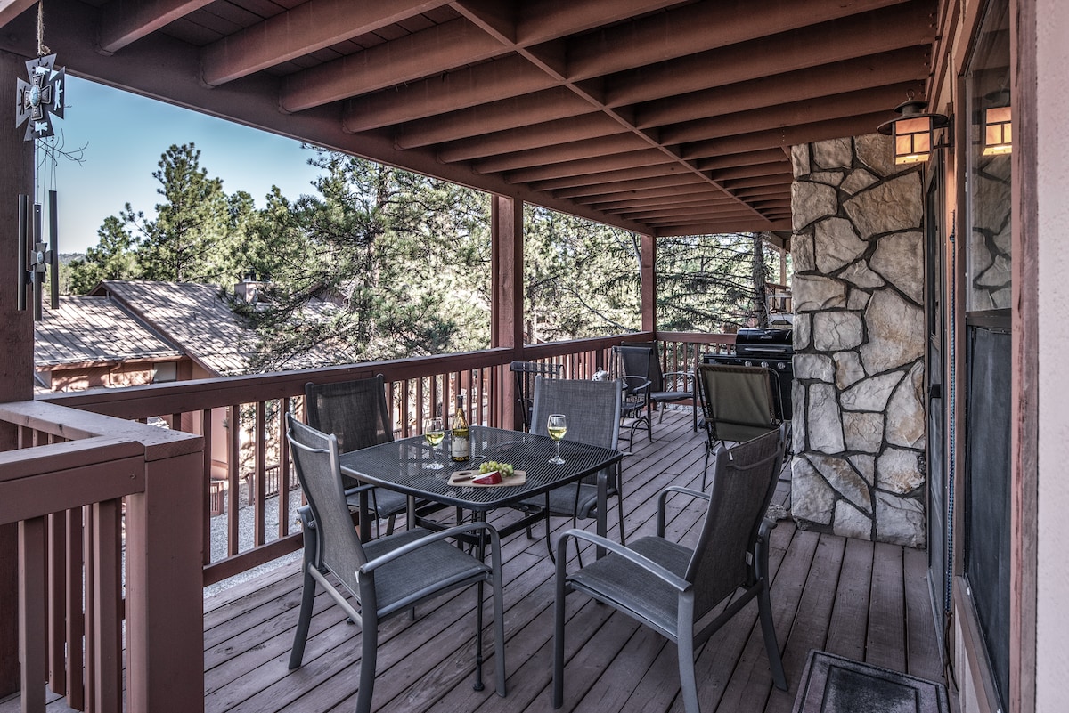 A spacious covered deck features a dining table surrounded by chairs, offering a relaxing outdoor space. A gas grill is positioned nearby, and the natural wooded surroundings provide a peaceful backdrop, inviting guests to enjoy meals and conversations in the fresh air.