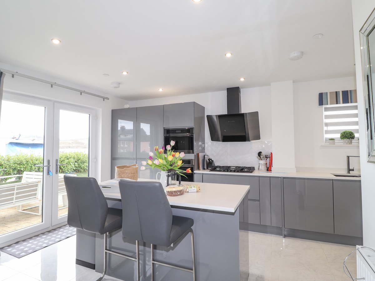 A modern kitchen space features sleek grey cabinetry and a central island with seating. Natural light enters through glass doors leading to an outdoor area. Fresh flowers are arranged on the counter, enhancing the clean and functional design of the space.