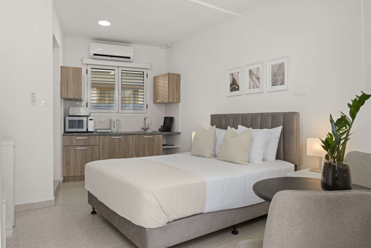 A modern studio layout is displayed, featuring a spacious bed with neutral bedding. Natural light filters through window blinds. A small kitchen area with essential appliances is visible in the background, complemented by wooden cabinetry. A cozy seating area includes a side table and a green plant.