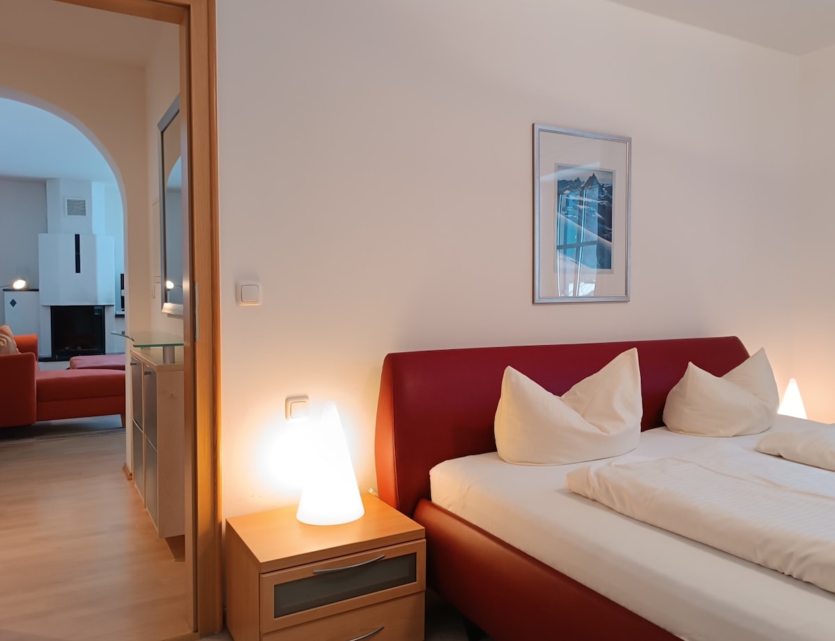 A spacious bedroom features a large bed with quilted bedding and two decorative pillows. Soft lighting emanates from bedside lamps, creating a calming atmosphere. A doorway leads to a living area, visible in the background, enhancing the open layout of the space.