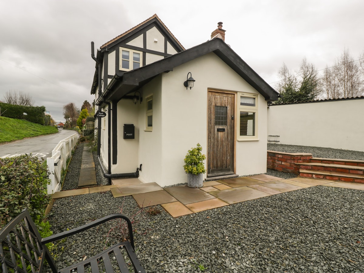 The exterior of the cottage presents a combination of traditional and modern architectural elements. A stone pathway leads to a front door framed by greenery, while seating is situated nearby on a gravel surface.