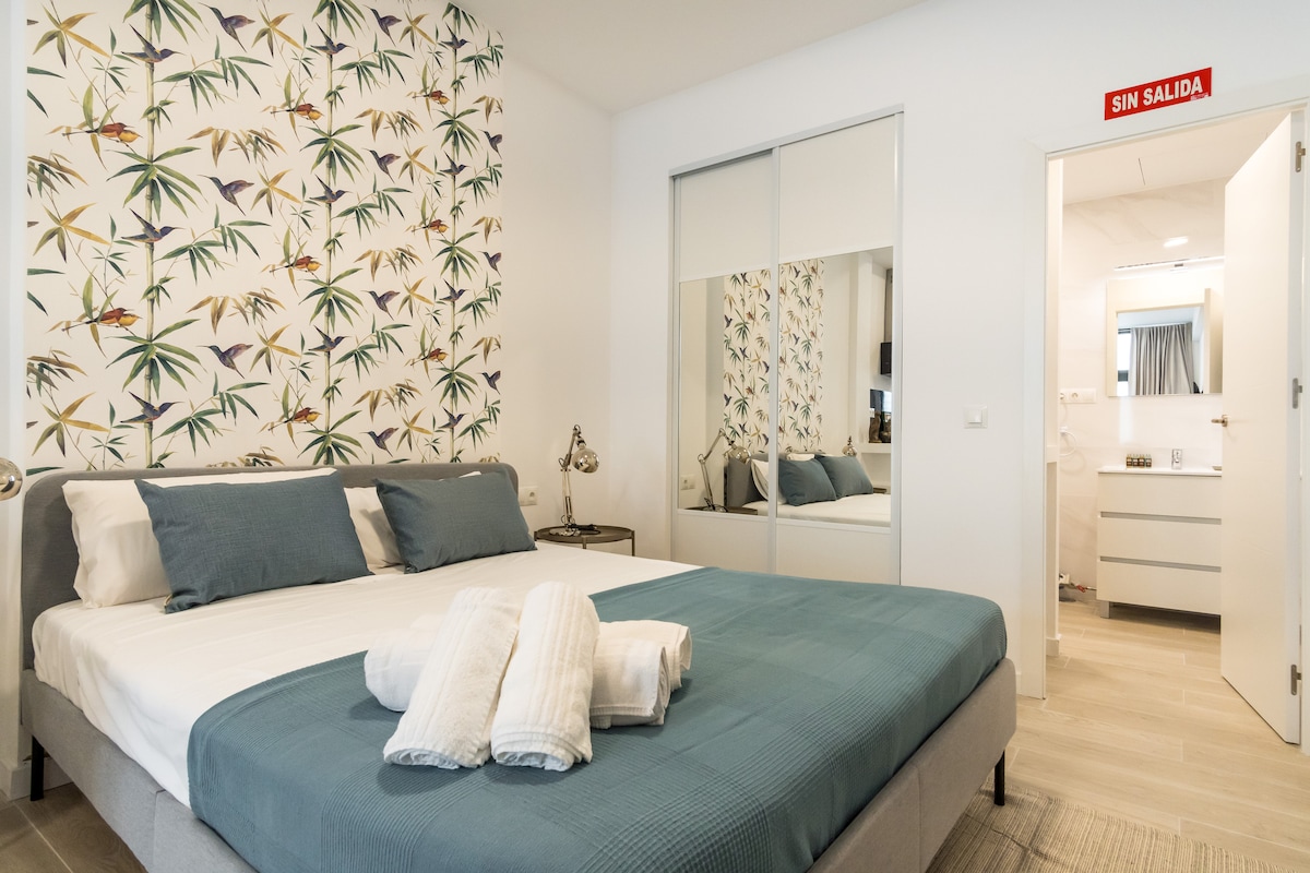 A modern bedroom is presented with a double bed covered in a light blue blanket. Two decorative pillows rest on the bed. A large mirrored wardrobe reflects the room, which features a tropical-themed accent wall. A doorway leads to the bathroom area in the background.