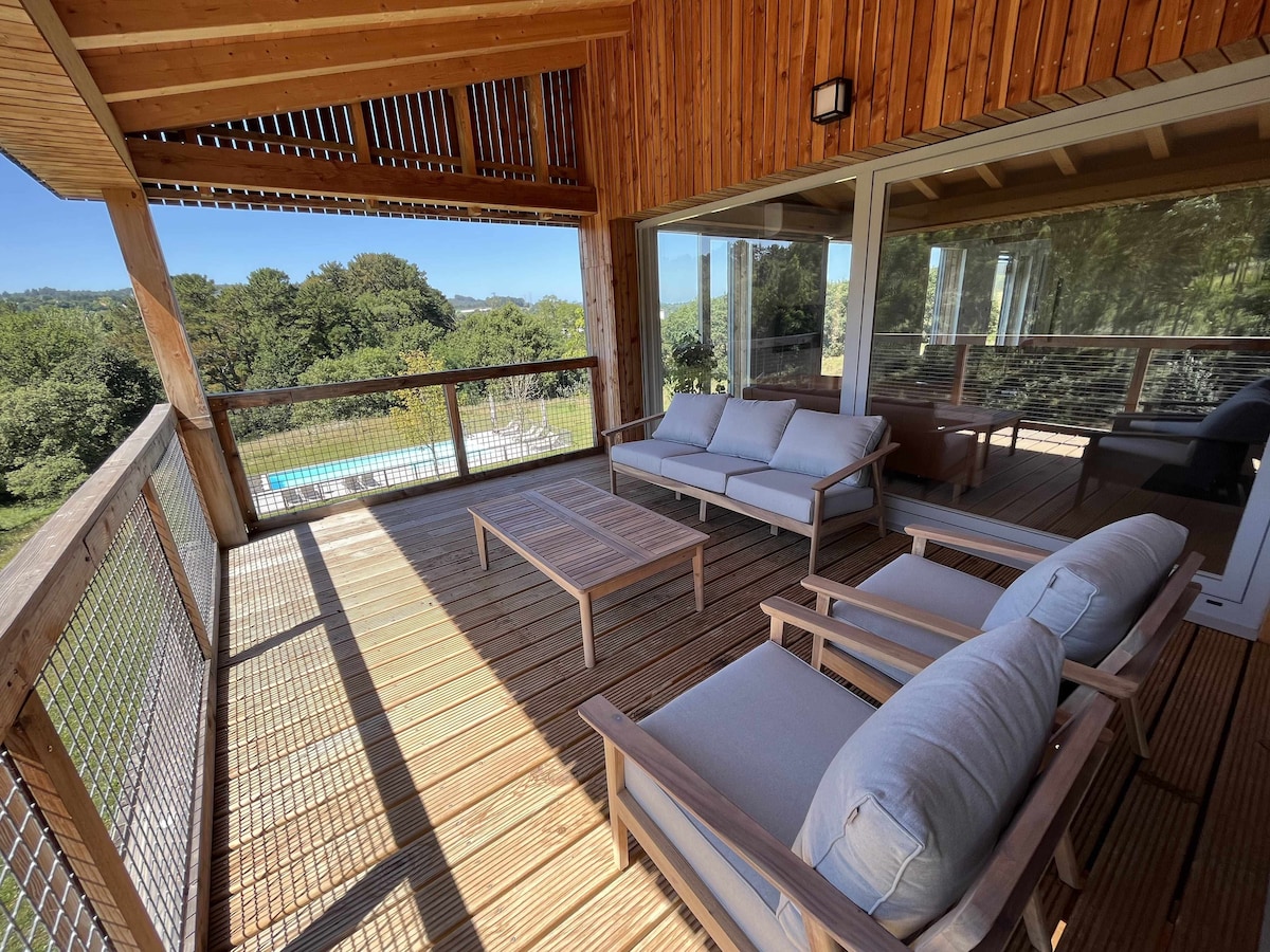 A spacious terrace is equipped with comfortable seating arrangements and a wooden coffee table. Large glass windows provide a view of the outdoor pool and surrounding greenery, allowing natural light to fill the space.