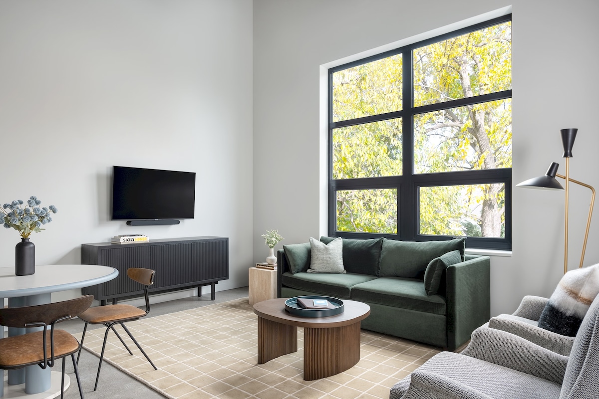 The living area features a dark green sofa and a light grey armchair, complemented by a round coffee table. A dining table with three chairs is positioned nearby. Large windows offer ample natural light, with views of foliage outside. A flat-screen TV is mounted on the wall.